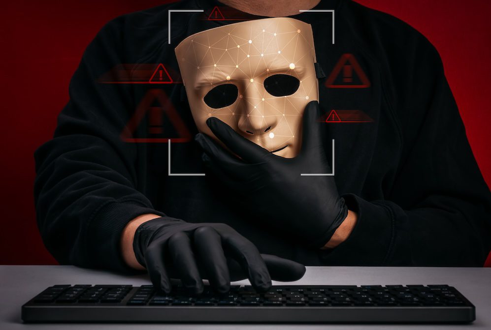 A person wearing black gloves holds a mask in front of their face, with a keyboard in front of them. Digital warning symbols, cybersecurity alerts, and facial recognition graphics are superimposed on the image.