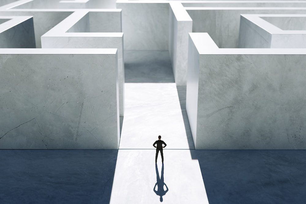 A person stands at the entrance of a large, white maze, casting a long shadow on the ground, appearing to contemplate which path to take—a minimalistic scene capturing the confusion often faced by growing businesses.