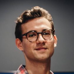 A young man with short brown hair and glasses is smiling slightly. He is wearing a plaid shirt and is photographed against a blurred gray background.