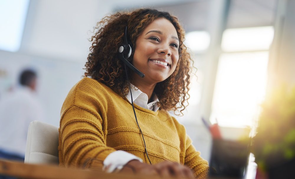 A woman with curly hair wearing a mustard yellow sweater and a headset smiles while working at a desk in a brightly lit office, showcasing the impact of AI in managed services.