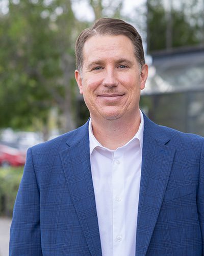 A man with short brown hair, wearing a blue checked suit jacket and white dress shirt, stands outdoors in front of blurred greenery and buildings, smiling at the camera with an air of executive leadership.