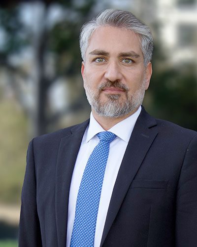 A man with gray hair and a beard, exuding executive leadership in a black suit, white shirt, and blue patterned tie, stands outdoors with a blurred background of trees and buildings.