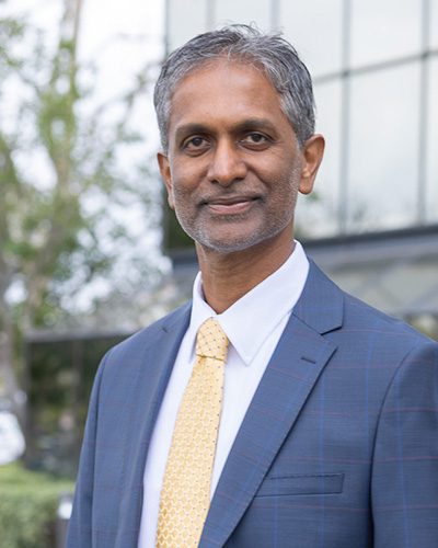 A middle-aged man with short gray hair and a trimmed beard, wearing a blue suit, white shirt, and yellow tie, stands outdoors in front of a modern glass building, reflecting confident executive leadership as he smiles slightly at the camera.