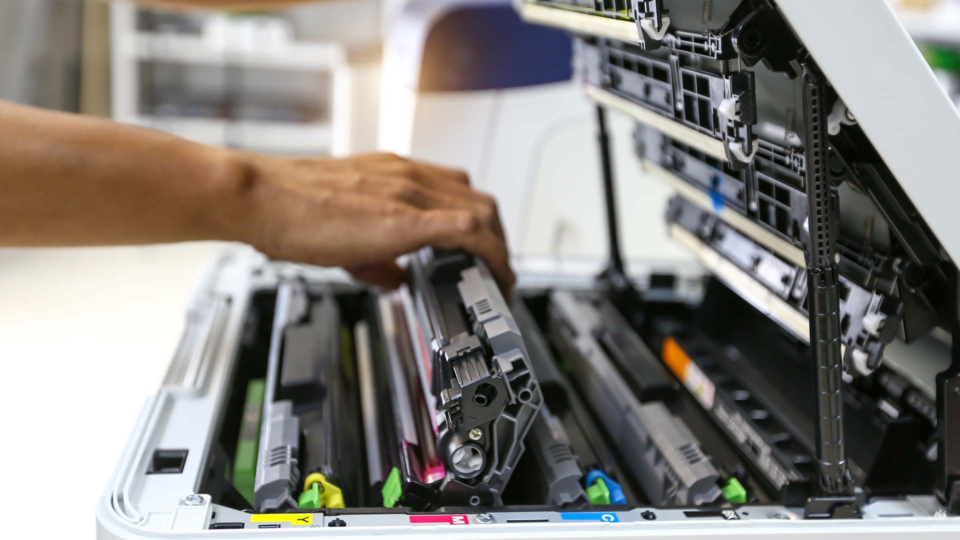 A person’s hand is installing or replacing a toner cartridge inside an open multi-function printer, with various colored toner slots visible—perfect for illustrating a Supplies Request or Supplies Order process.