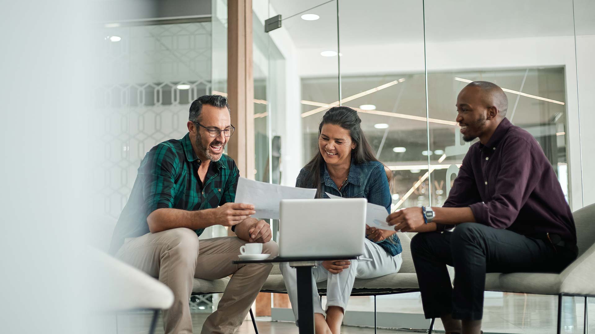 Three people sit together in a modern office, smiling and reviewing papers labeled Sales Request in front of a laptop on a small table, suggesting a collaborative and positive work meeting.