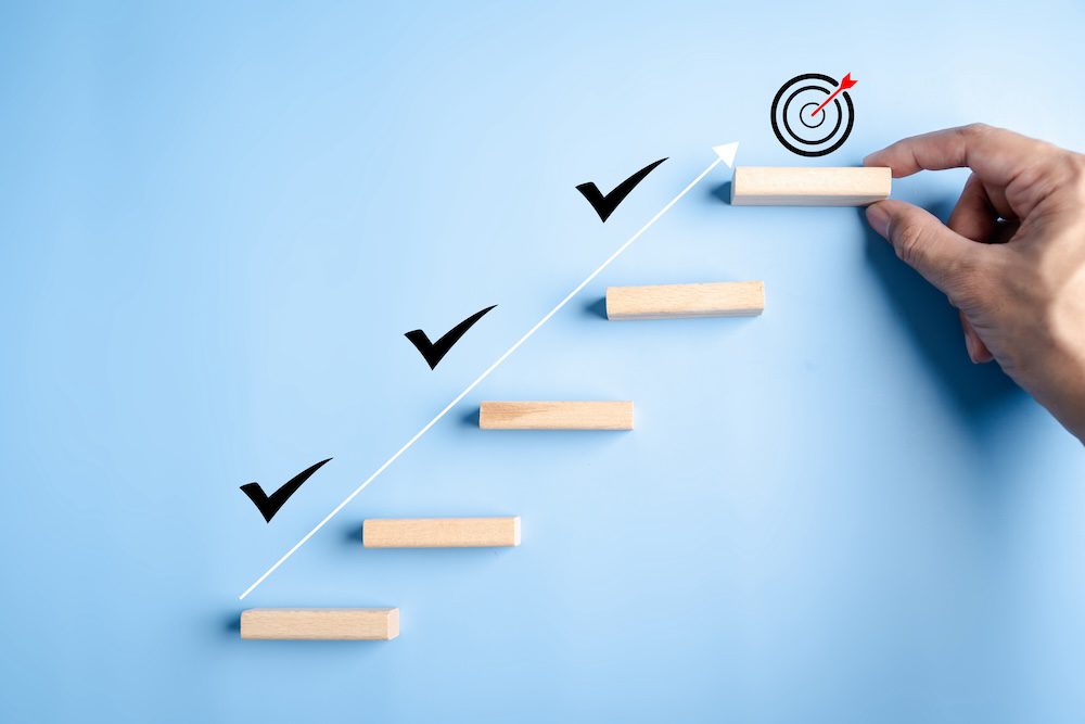 A hand places the final wooden block of a staircase ascending along a white arrow with check marks, leading to a target—symbolizing progress in closing the IT skills gap and achieving goals through effective IT training.