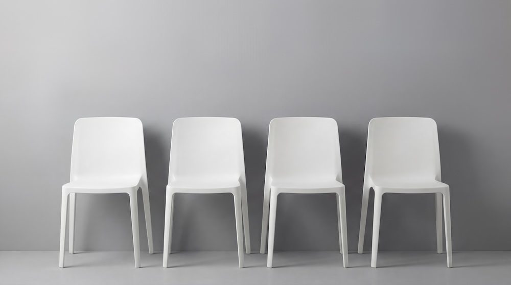 Four identical white plastic chairs are evenly spaced in a row against a plain light gray wall and floor, subtly reflecting the emptiness and impact of the IT workforce shortage in a minimalist, modern scene.