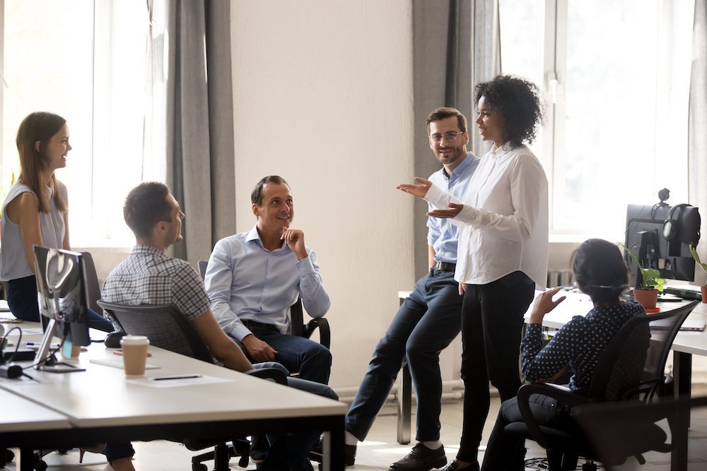 A group of five people in business casual attire have a discussion in a bright office. One woman stands and speaks, sharing key questions to ask your IT team while the others sit, smiling and engaged. Desks, computers, and windows are visible.