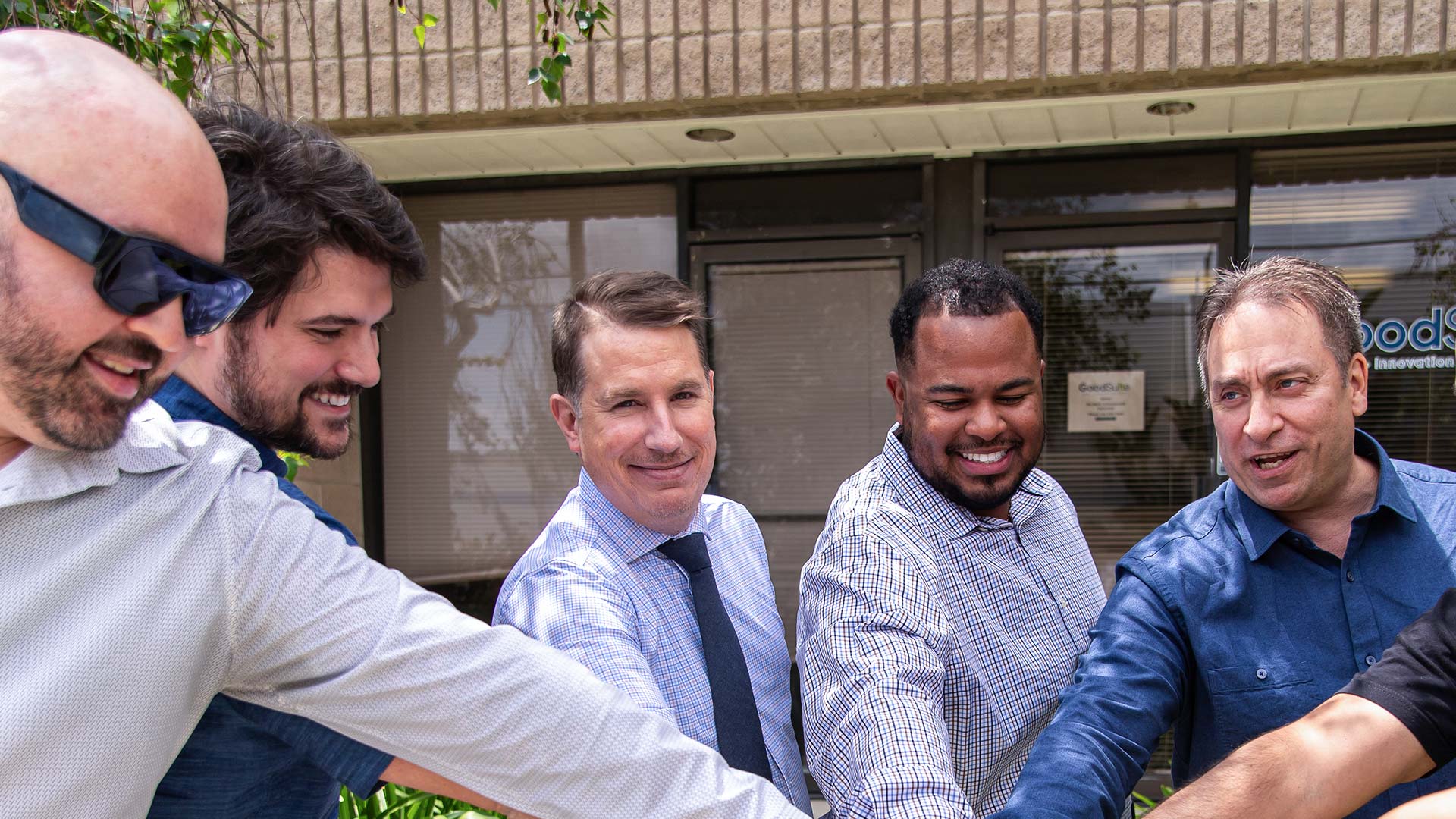 Five men stand in a line outdoors with their hands stacked together in the center, smiling and appearing unified. They are dressed in business casual attire, and a building with windows is visible in the background.